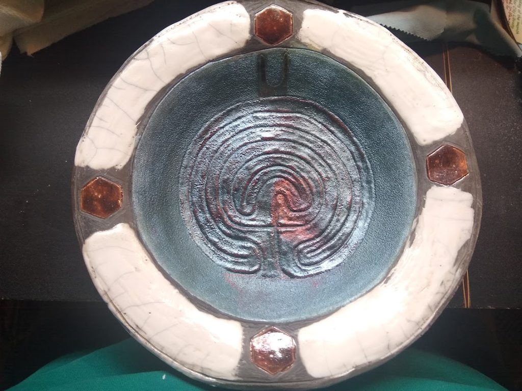 raku fired ceramic dish, the outer edge is glazed white and divided by four copper colored hexagons at the 12, 3, 6 and 9 positions, the center of the dish has an inscribed Cretan labyrinth design with an incised stylized horns (known as the horns of consecration) above the labyrinth and under the top copper hexagon).