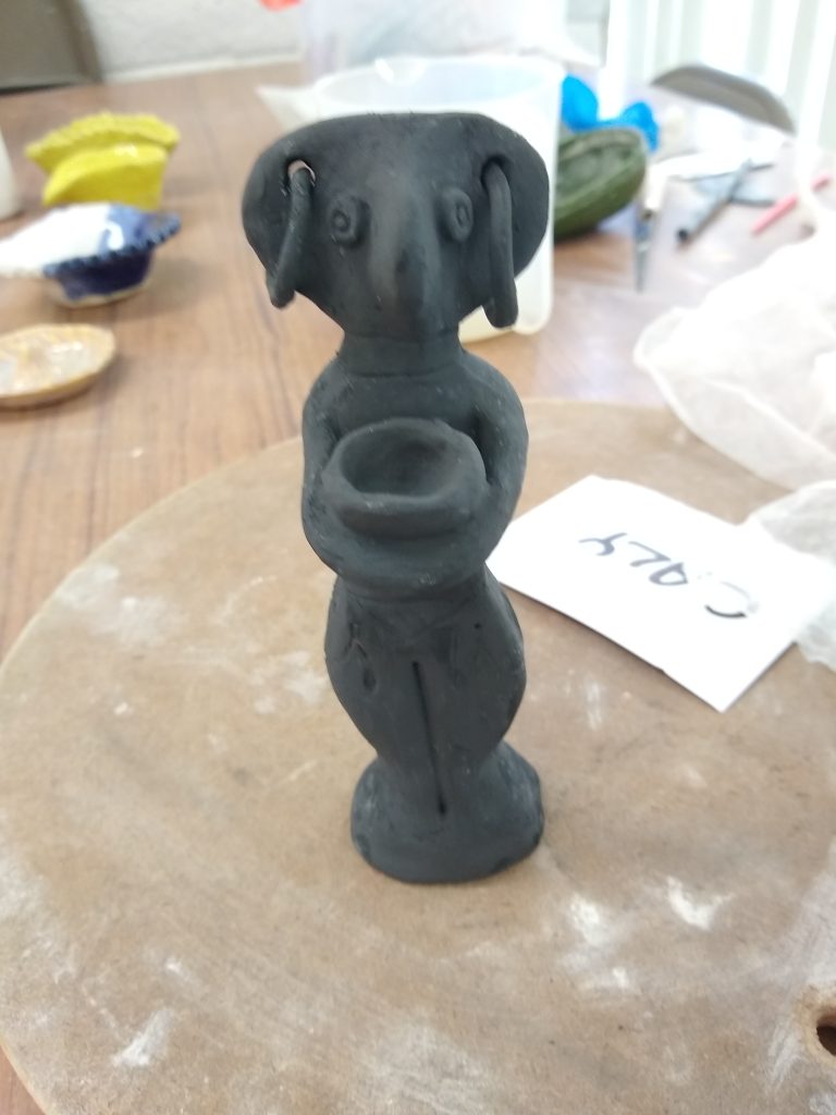 greenware clay figurine of a beaked female body with two large ear rings holding a bowl covered in black oxide