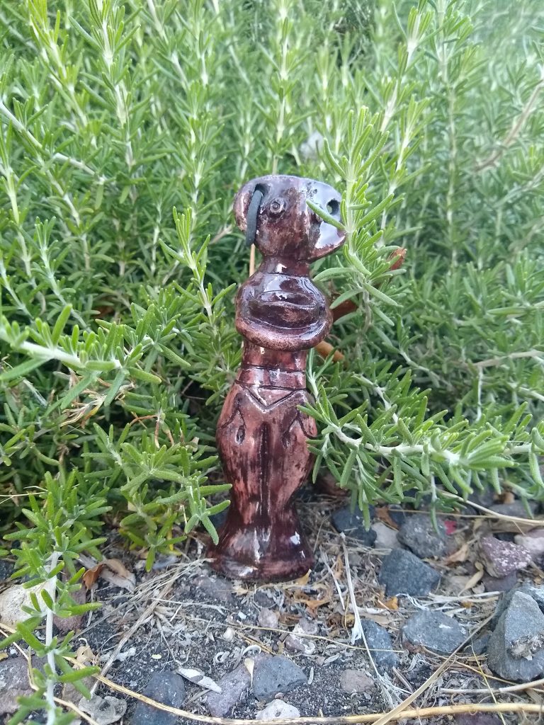 fired clay figurine of a beaked female body with two large ear rings holding a bowl, it now is glossy with dark reddish to black coloring, one earring is missing, it is positioned on the ground nestled into rosemary branches